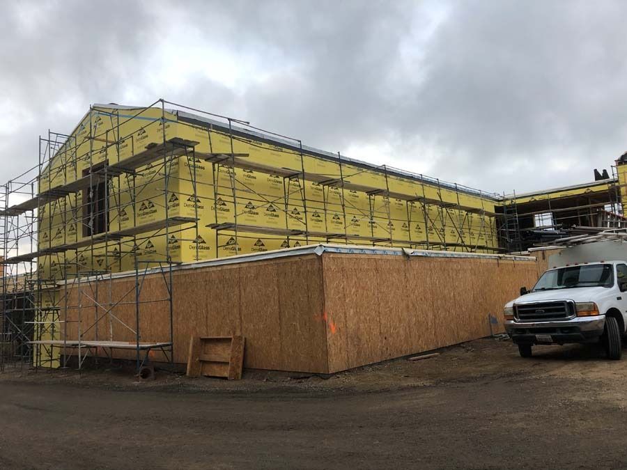 Building under construction, exterior covered in yellow insulation, scaffolding and brown paneling. Cloudy sky, pickup truck.