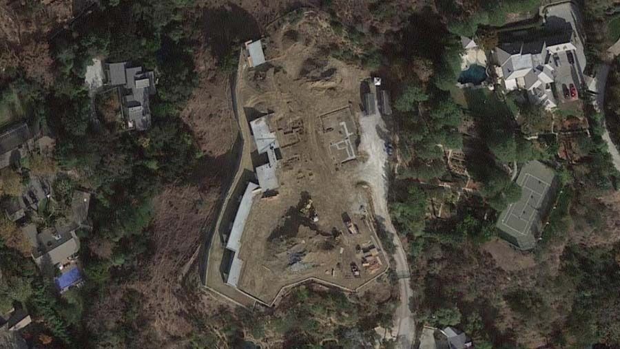 Aerial view of a construction site surrounded by trees and houses. Gray buildings and dirt path visible.