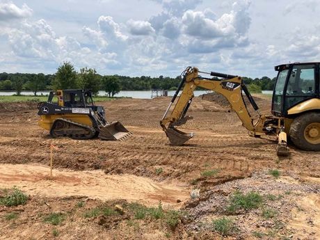 Two yellow construction vehicles on a dirt site, a backhoe and a skid steer, under a cloudy sky.
