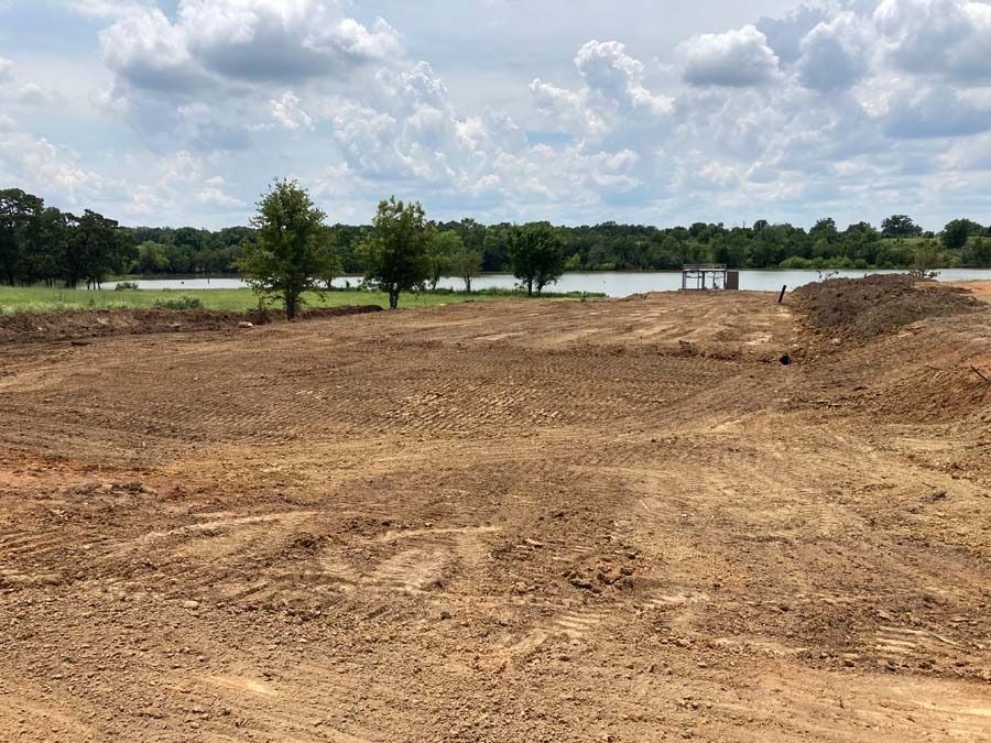 Brown dirt lot in front of lake, with trees and cloudy sky in the background.