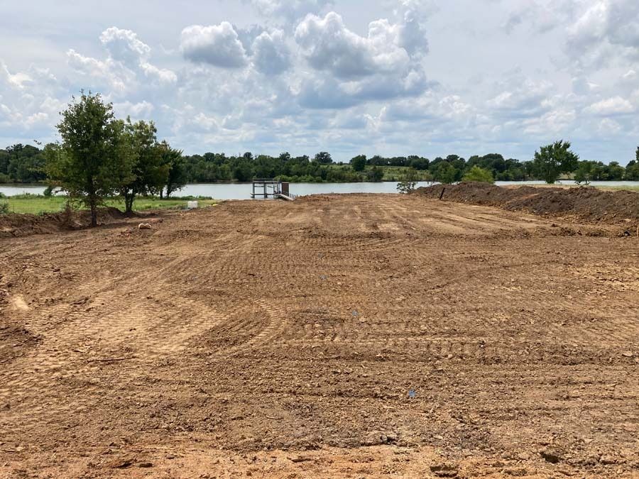 Brown dirt lot slopes down to a body of water under a cloudy sky, near green trees.