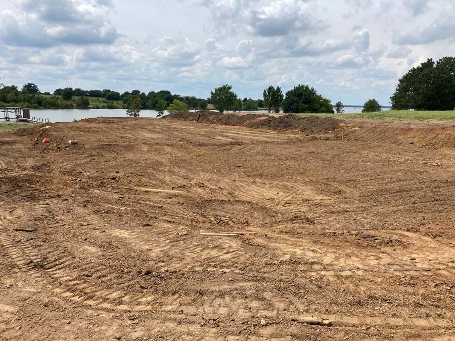 Brown dirt field under a cloudy sky, with water and trees in the background.