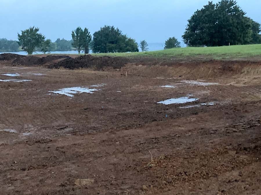 Muddy, freshly dug construction site with puddles; trees and water in the background.