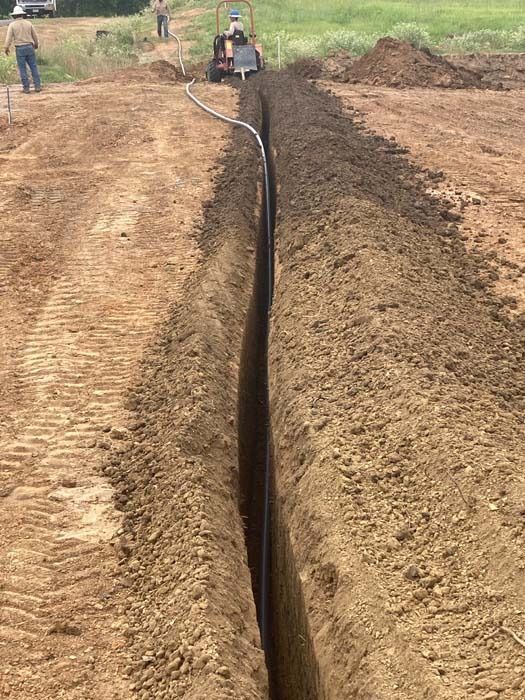 Trench dug in a field with a black pipe, possibly for utilities, being laid by a machine.