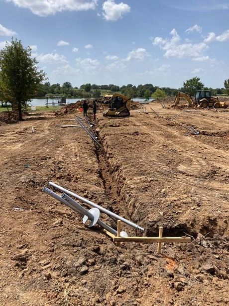Construction site with trench, equipment, and workers in foreground. Lake in the background. Sunny day.