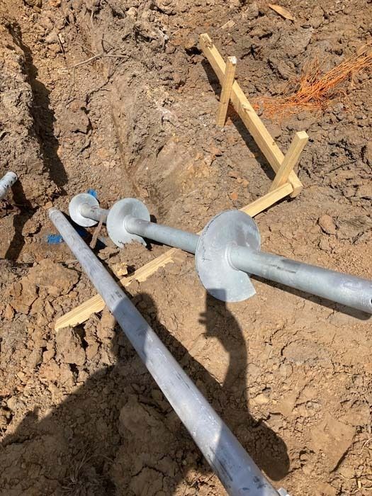 Metal helical piers installed in a dirt trench, used for foundation support; wooden guides in the background.