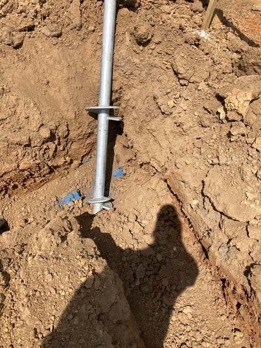 A metal ground screw installed in brown soil, with a shadow cast over the scene.