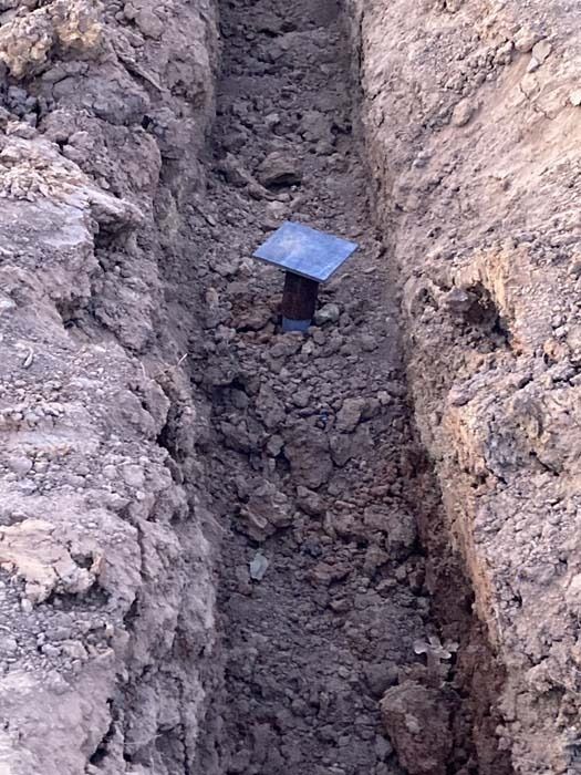 A narrow trench dug in the dirt, with a metal post base visible in the center.