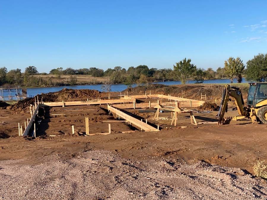 Construction site: wooden frame of a building foundation. A small excavator is present. Background has a lake and trees.