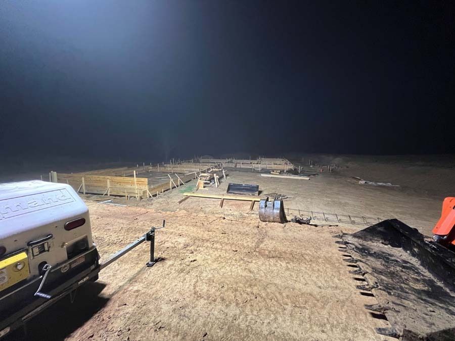 Nighttime construction site with dirt, equipment, and temporary fencing under a dark sky.