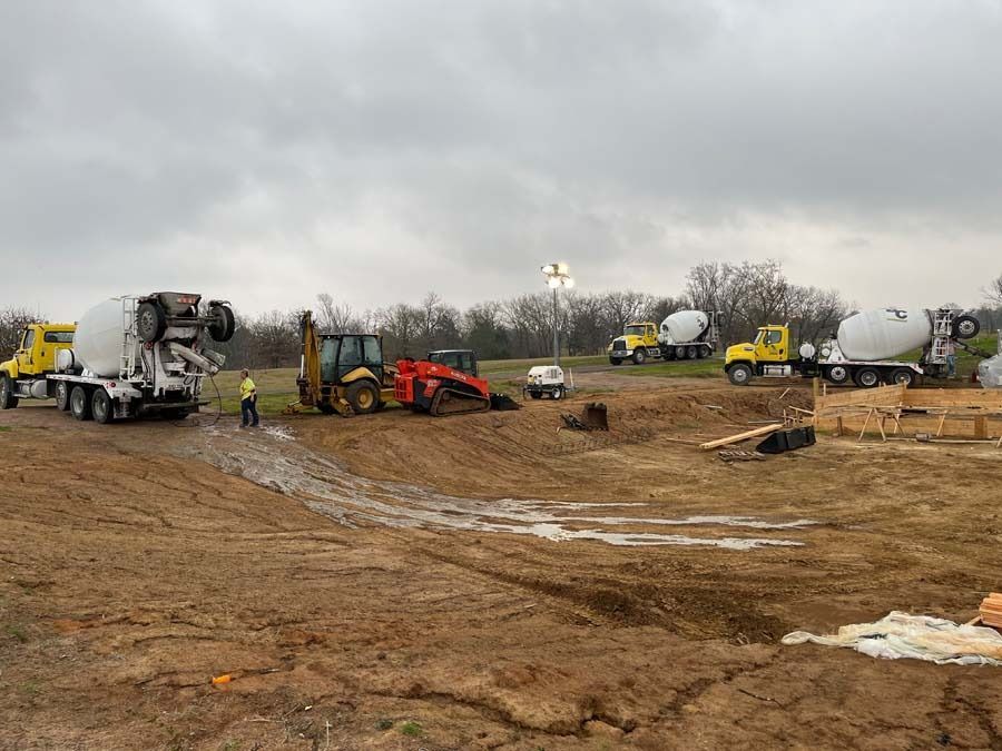 Construction site with cement trucks, a backhoe, and workers under a cloudy sky.