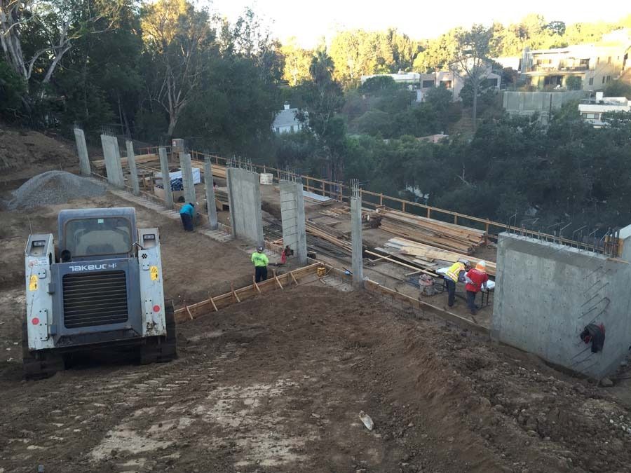 Construction site with workers, concrete forms, and a skid steer on a hillside.