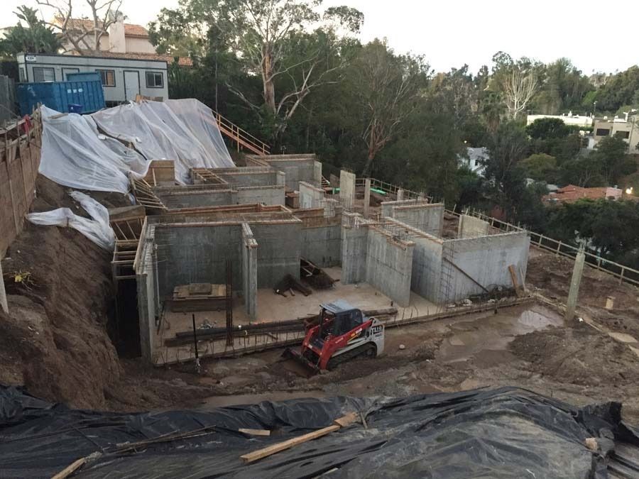 Construction site on a hillside with concrete foundations, a small loader, and tarps covering dirt.