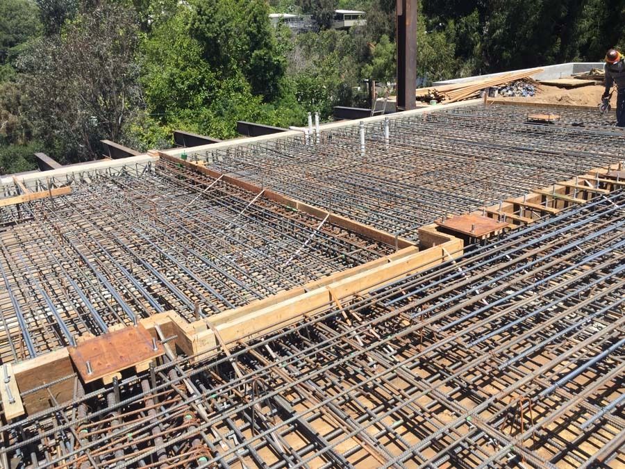 Construction site: rebar grid laid on wooden forms, ready for concrete, surrounded by trees.