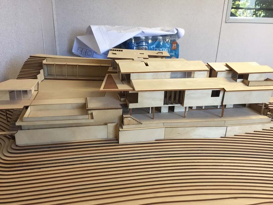 Architectural model of a multi-level wooden house on a topographical base, with blueprints and water bottles in the background.