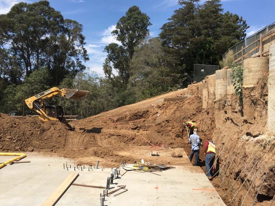 Construction site: Excavator on a dirt hill, workers near retaining wall, concrete foundation.