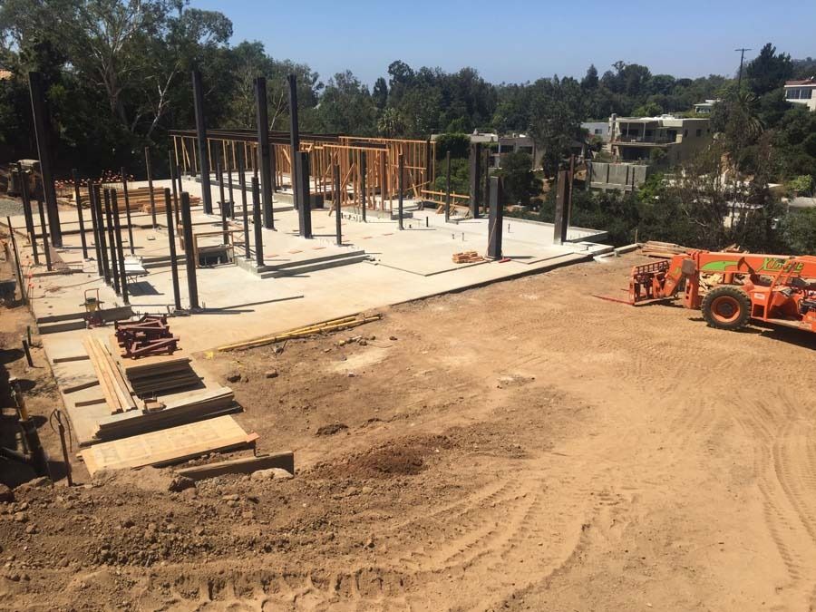 Construction site with steel beams, wood framing, and concrete slab, set against a hillside with trees and buildings.