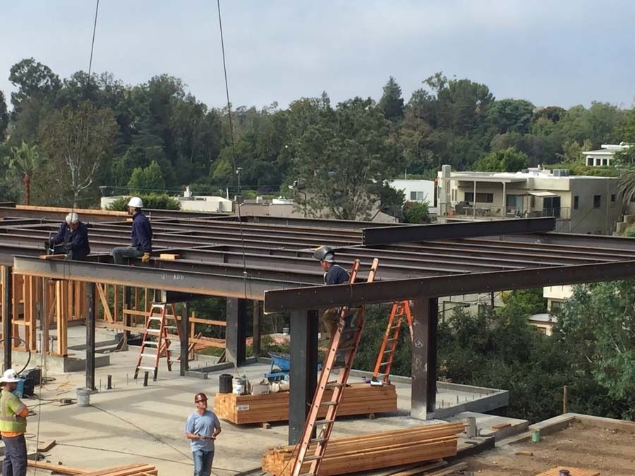 Construction workers installing steel beams on a partially built structure, outdoors with trees and buildings in the background.