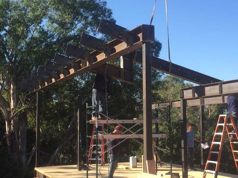 Construction workers assembling a steel beam structure outdoors; several are on ladders and scaffolding.