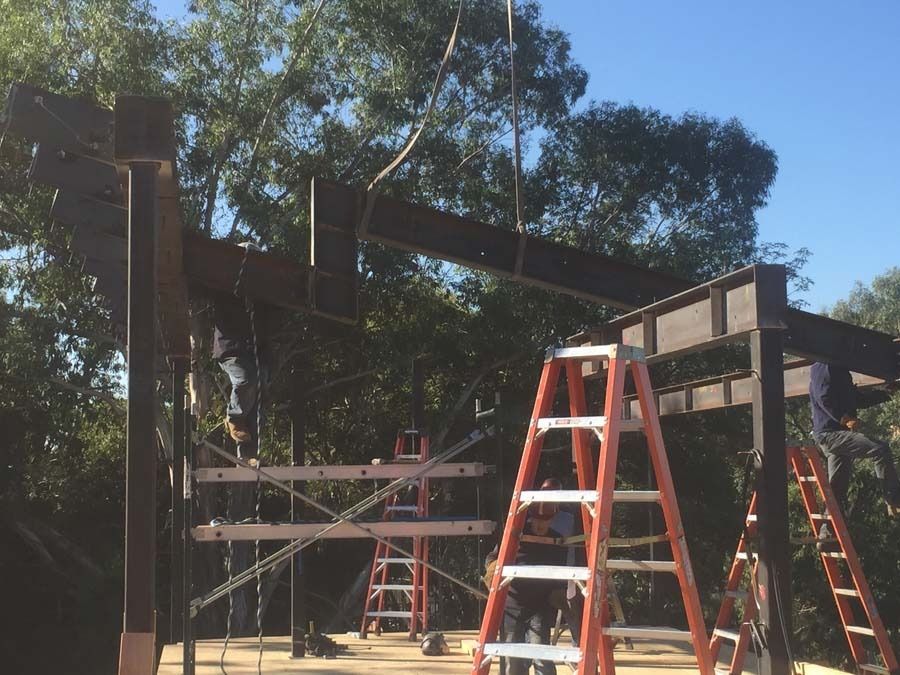 Construction workers assembling steel beams on a sunny day. Ladders, trees, and blue sky visible.