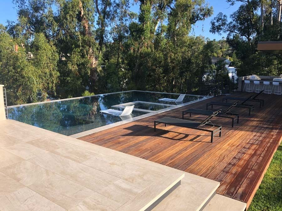 Wooden deck and infinity pool surrounded by trees, with lounge chairs and a sunny sky.