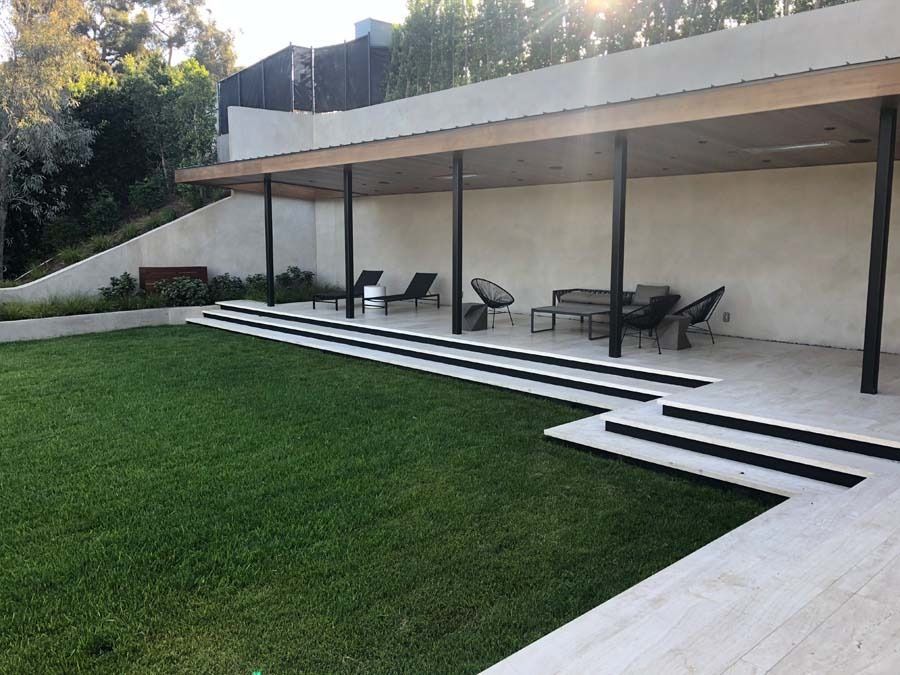Lawn with stone steps leading to a covered patio area with furniture. Sunlight filters through the trees.