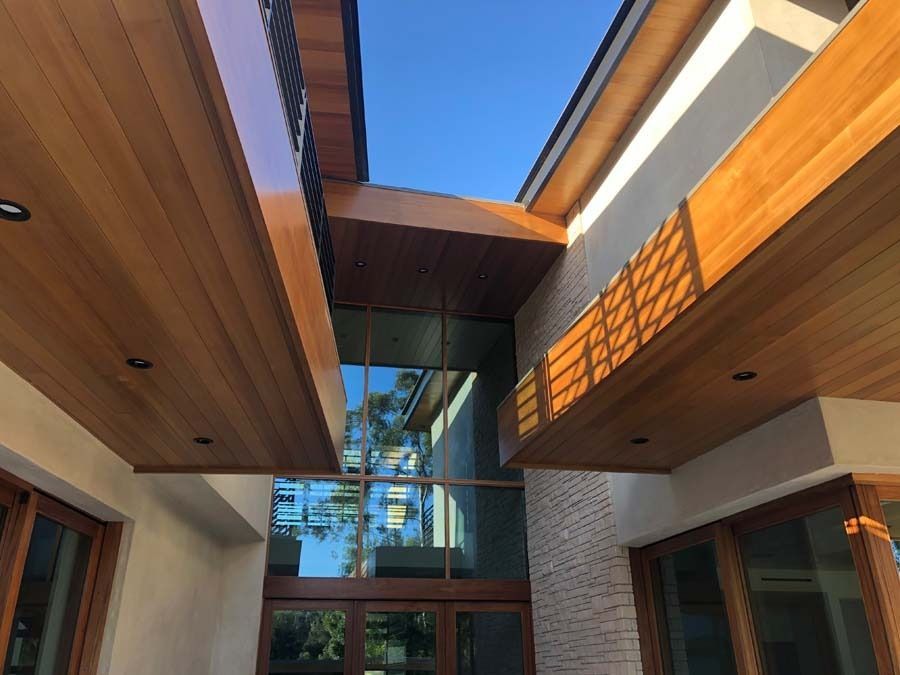 Wooden soffits and beams frame the glass entry of a modern home, against a clear blue sky.