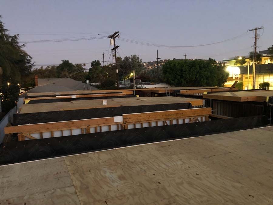 Construction of a flat roof with exposed wooden beams, black underlayment, and white supports. Dusk setting.