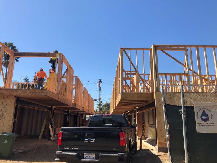 Construction site: two wood-framed buildings under construction, workers, a black truck, and a blue sky.