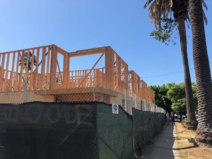 Construction site with wooden framing, behind a chain-link fence. Palm trees and sidewalk. Blue sky.