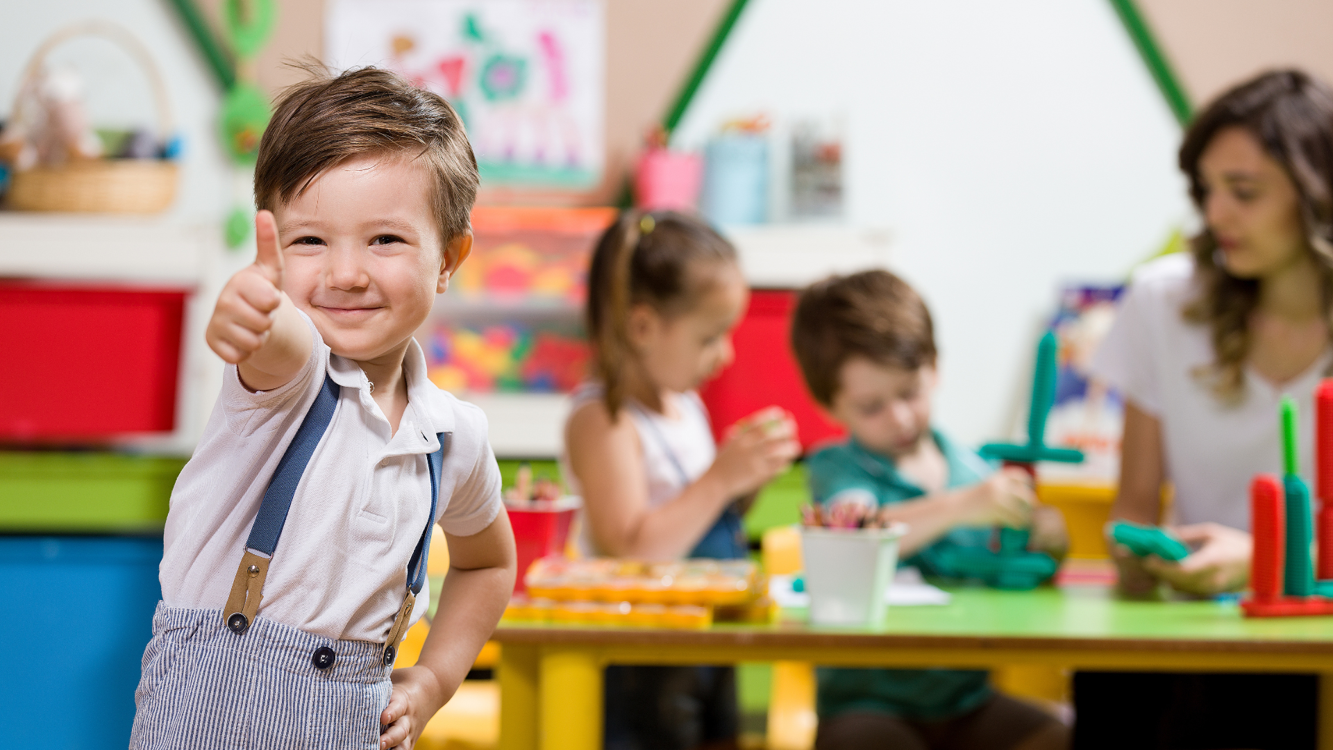 Boy with thumb up, smiling in a classroom with other children and a teacher.