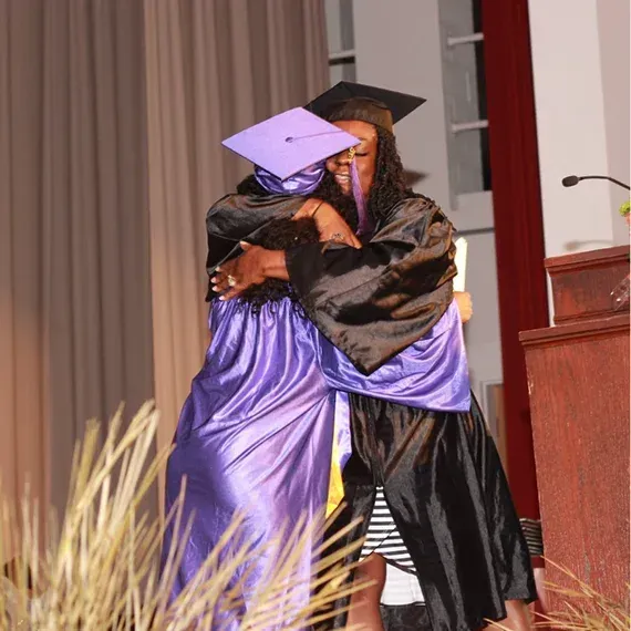 Two graduates in caps and gowns embrace warmly on a stage.