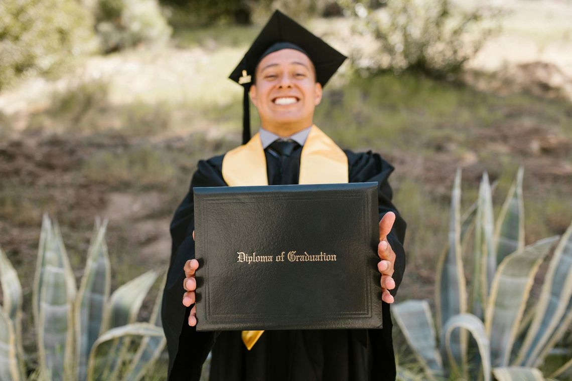 A smiling graduate wearing a cap and gown holds a diploma cover toward the camera against a background of desert plants.