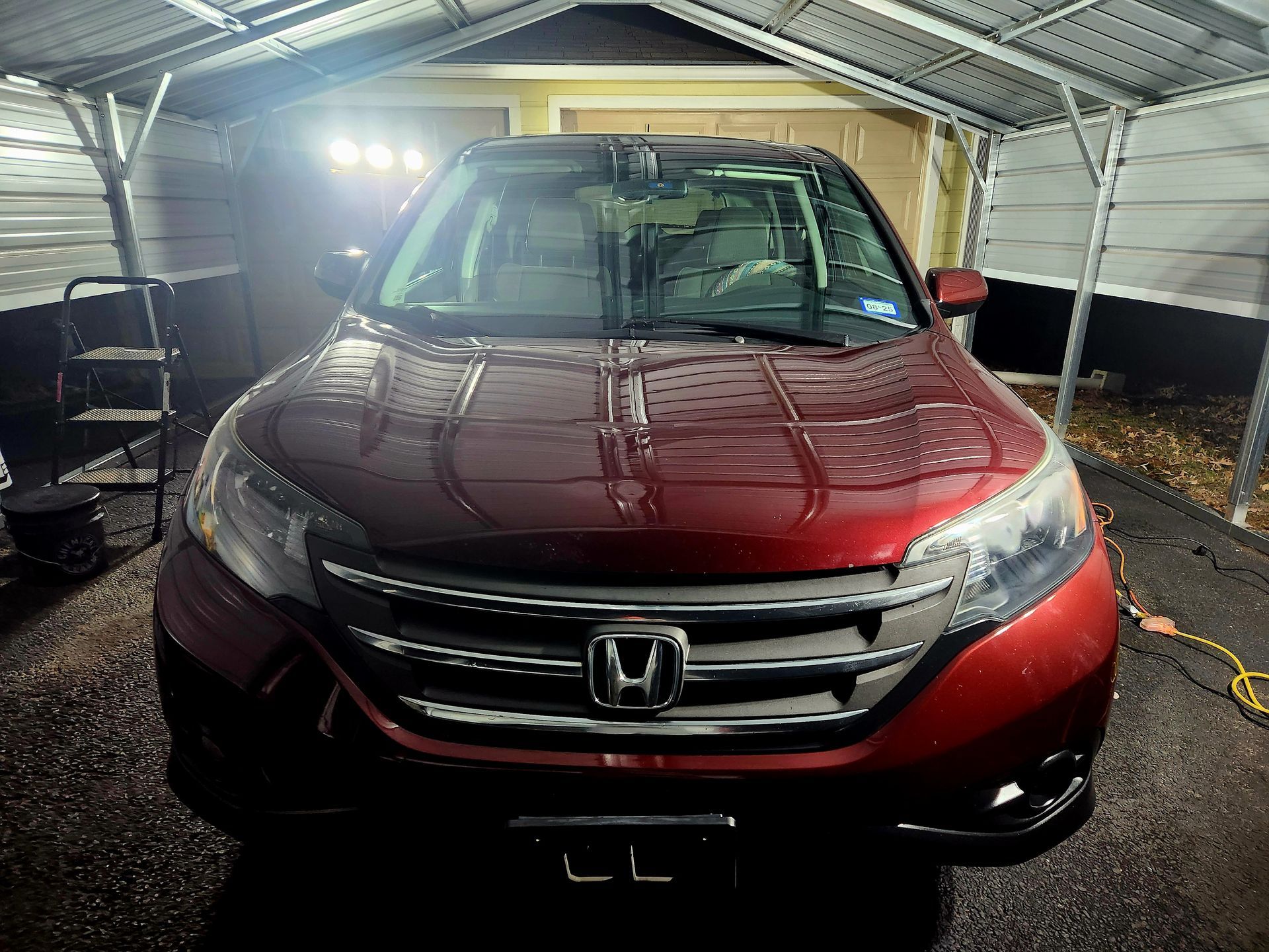 Red Honda CRV parked under a carport; front view; wet windshield, bright lights.