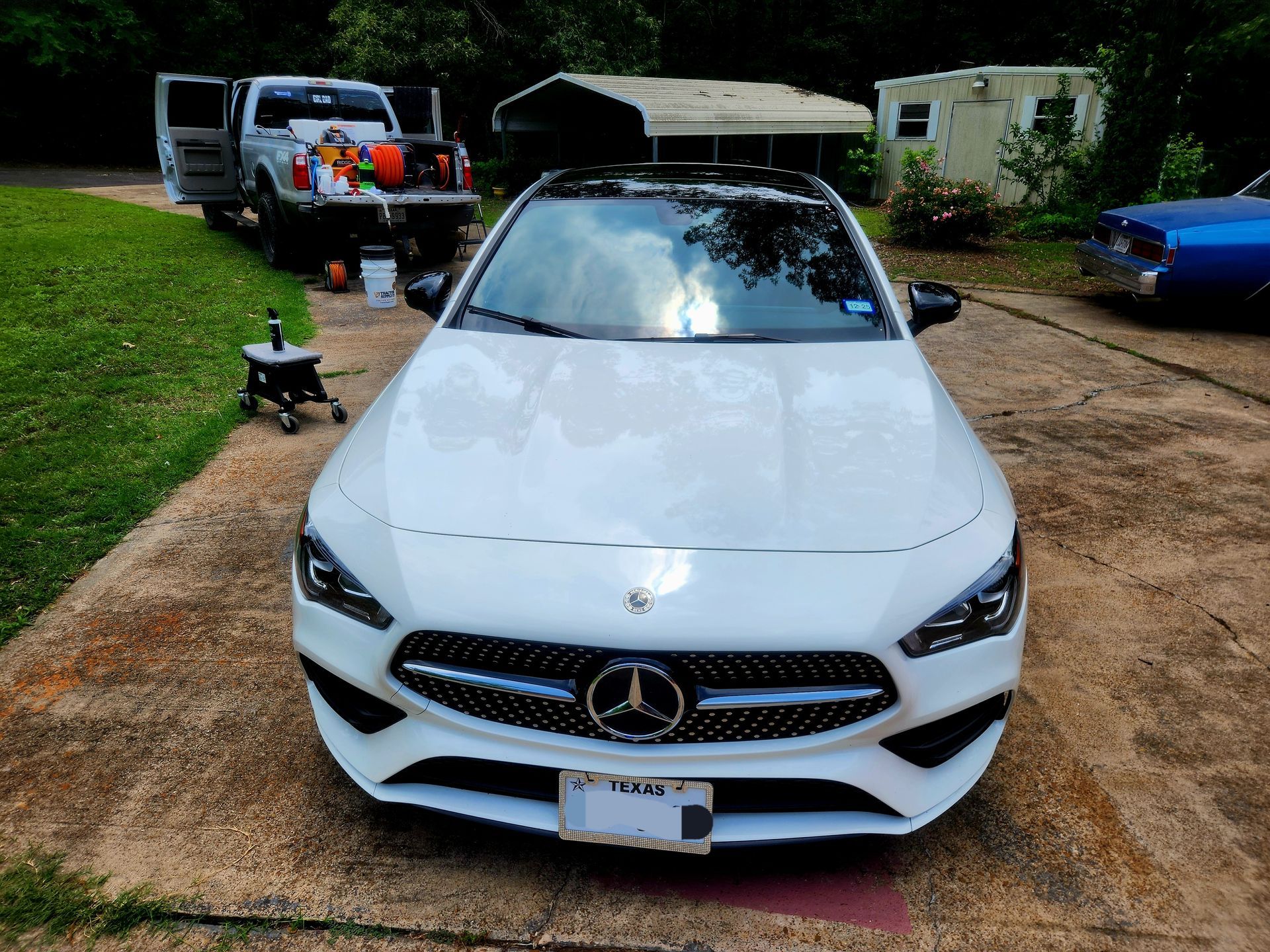 White Mercedes-Benz car parked in driveway. White truck with open doors in background.
