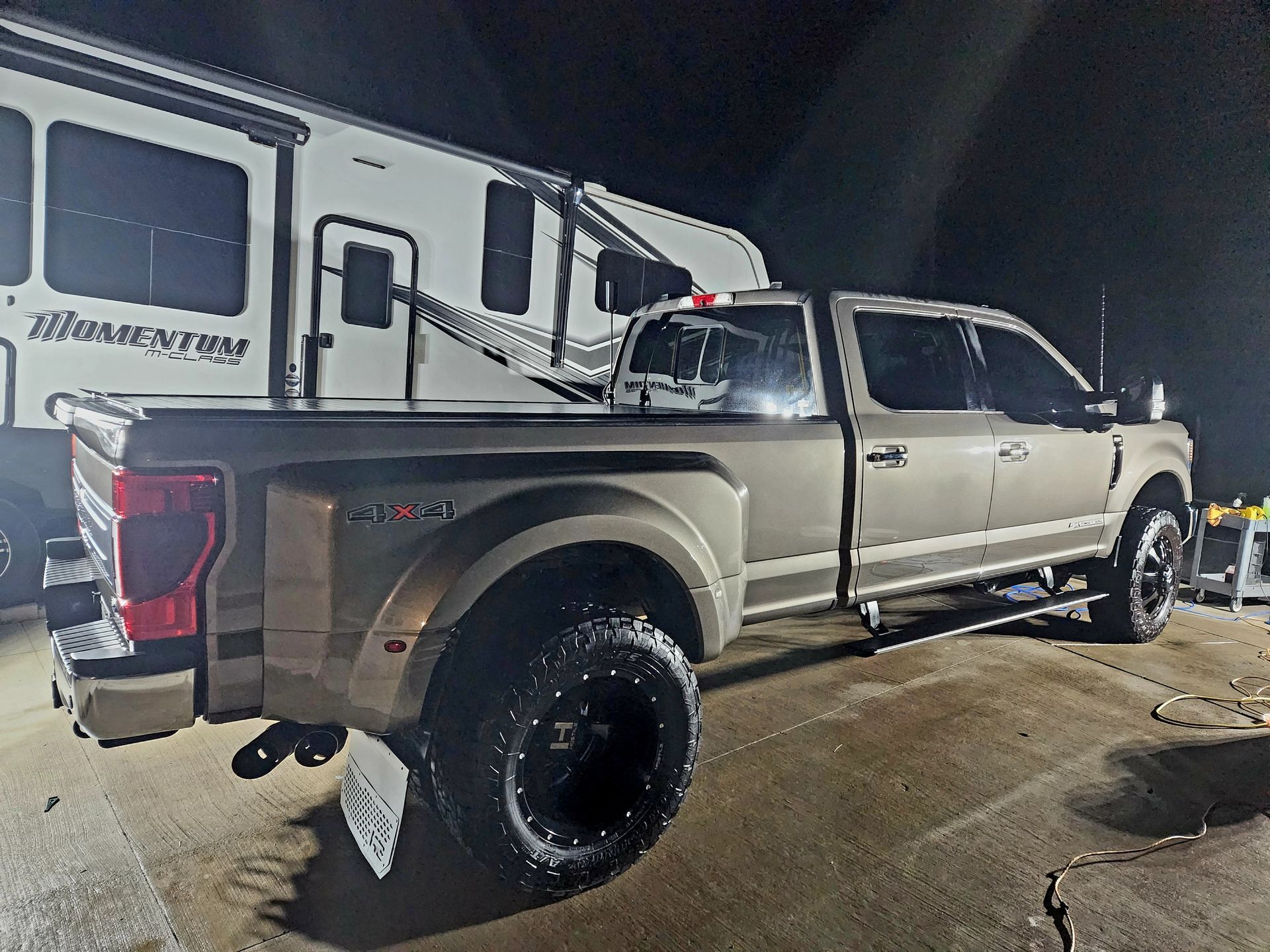 Tan Ford dually truck parked near a camping trailer at night. Black rims, 4x4 decal, and license plate visible.