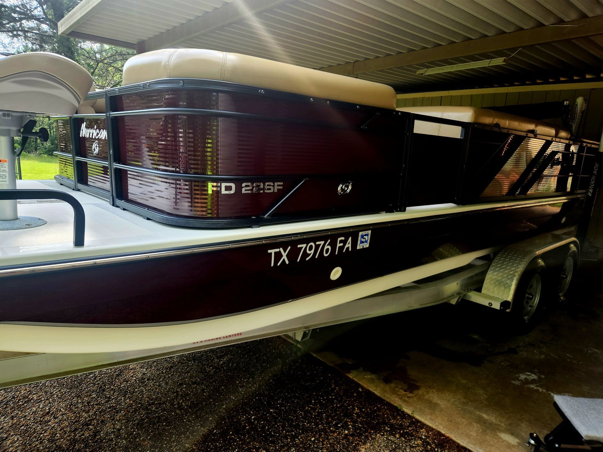 A maroon pontoon boat on a trailer, under a carport. The boat has a beige canopy, and white deck.
