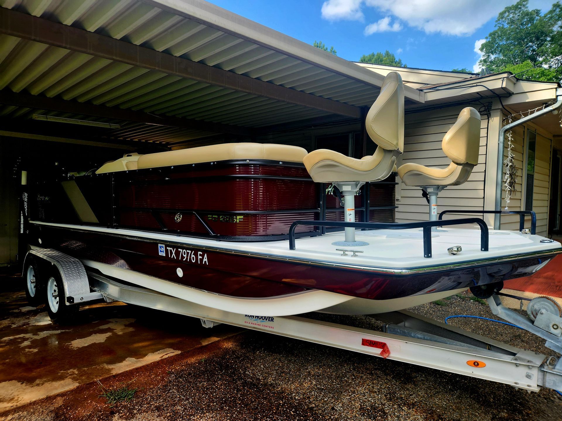 Boat on a trailer under a carport, burgundy and white, two beige fishing chairs, Florida registration.