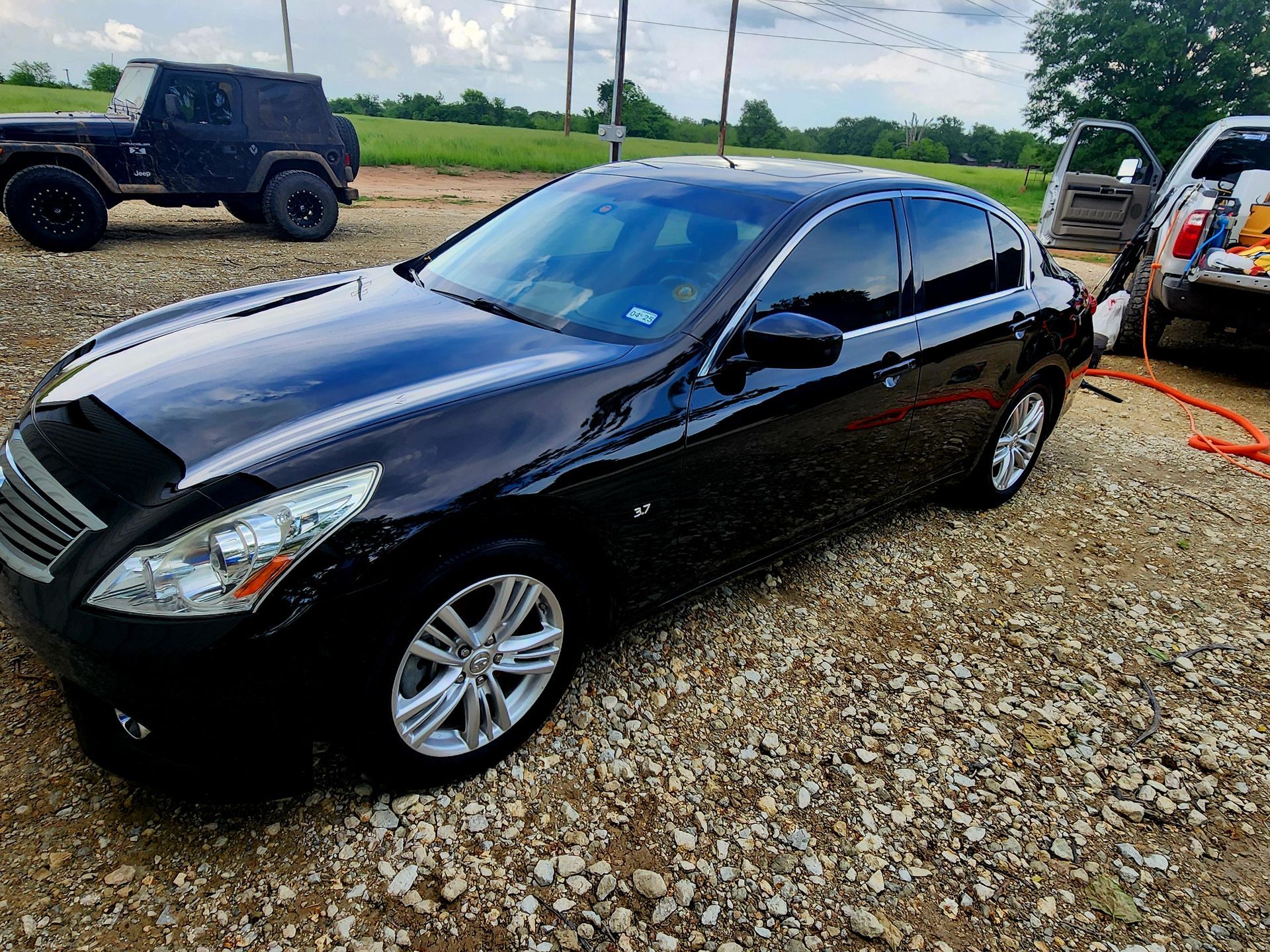 Black Infiniti sedan parked on gravel; a Jeep and a van in the background.