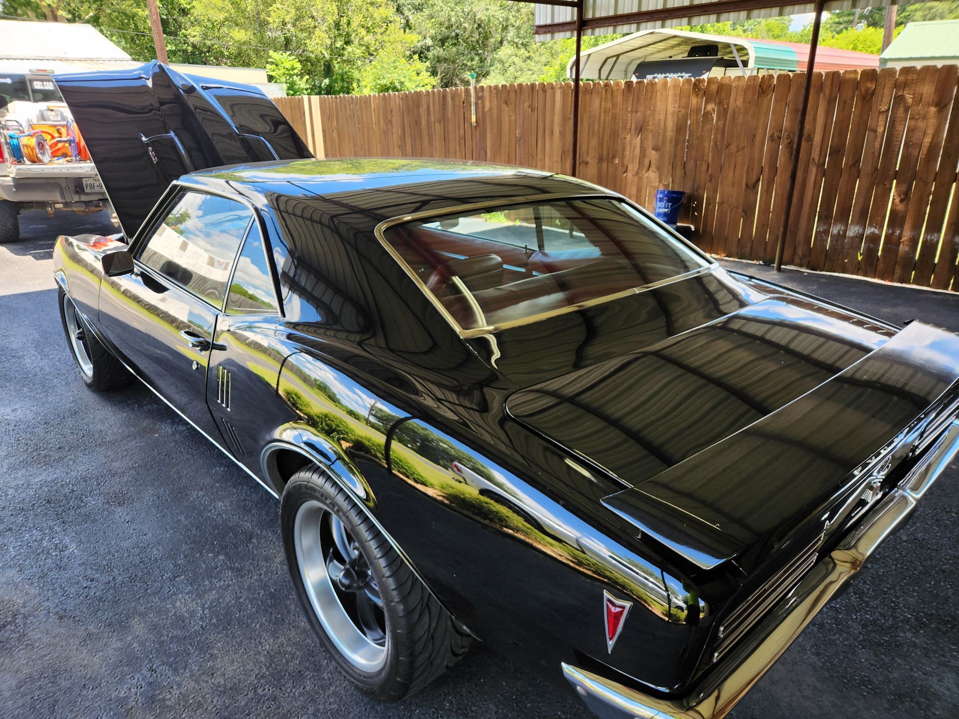 Black Pontiac Firebird with hood open, parked outside near a wooden fence.