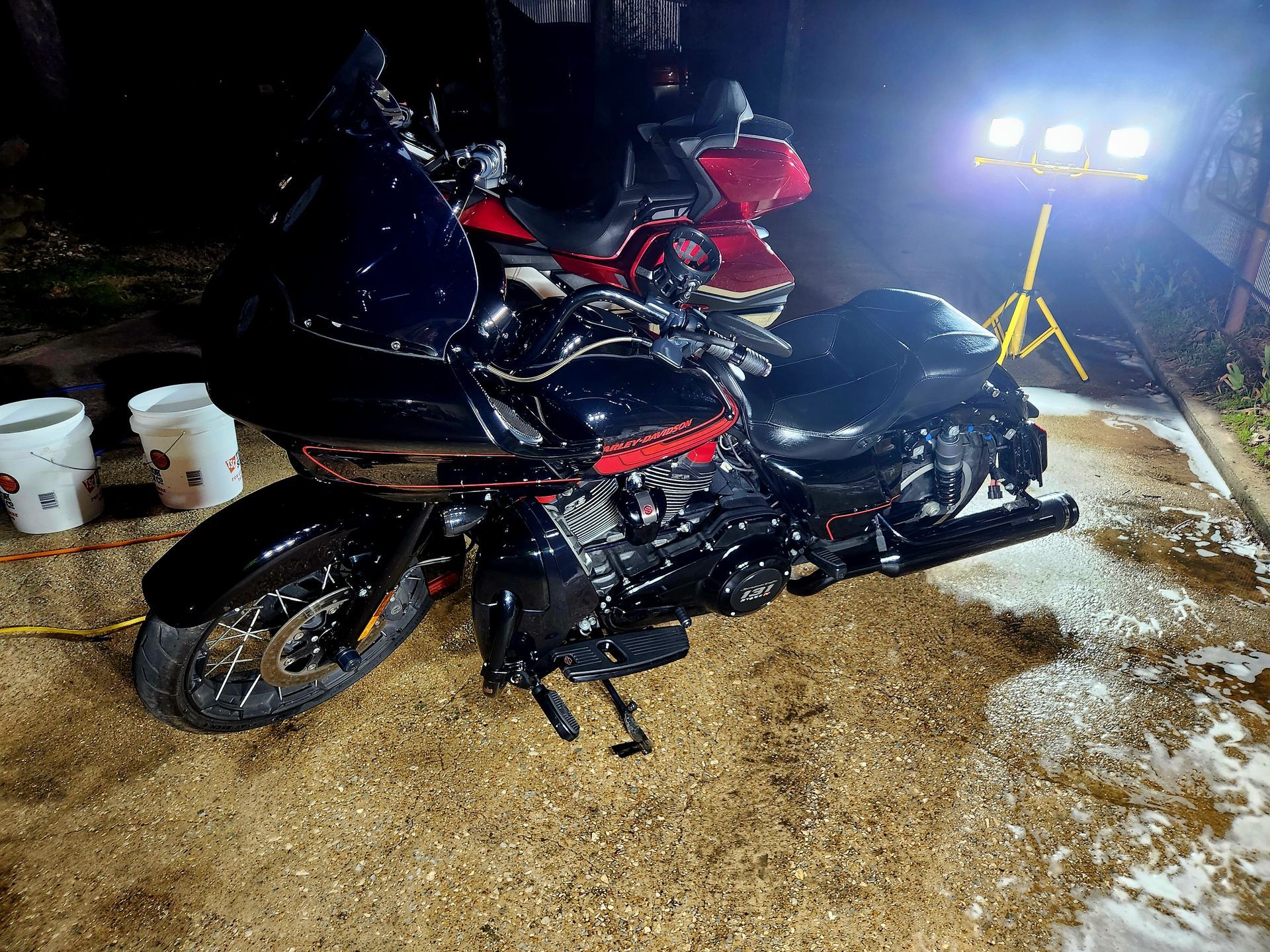 Motorcycles being washed outdoors at night, black and red bikes with foam and bright lights.