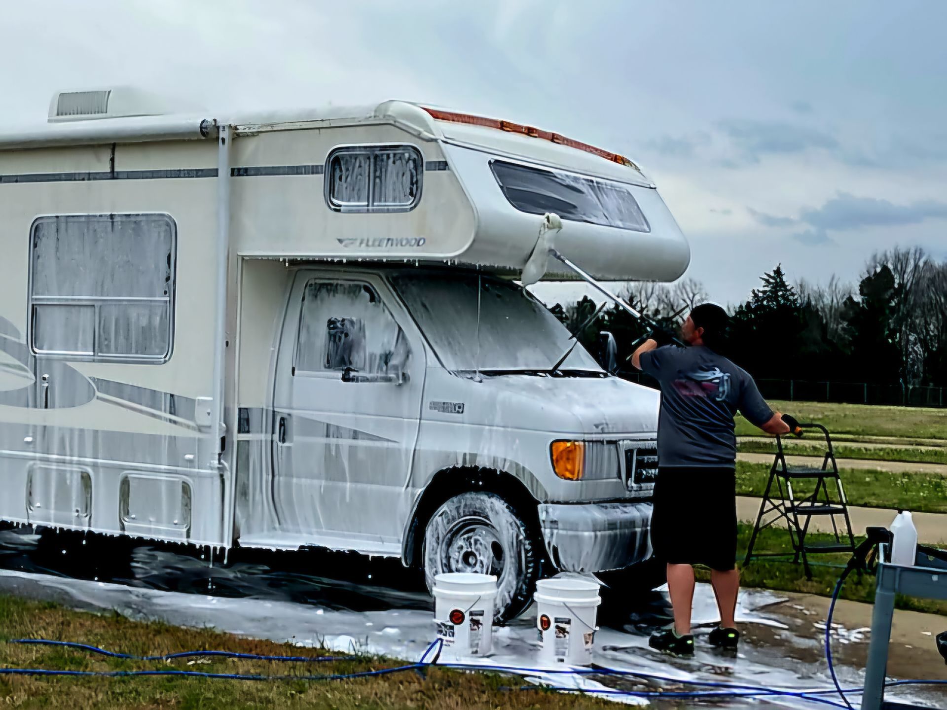 Man washing a white RV with foamy soap outdoors, using a ladder and buckets.