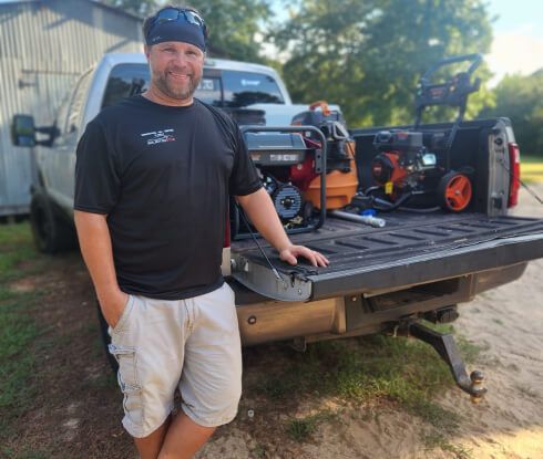 Man in cap stands by truck with power washers in the bed, smiling. Outdoors.