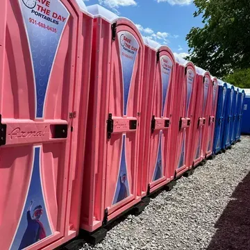 A row of pink and blue portable toilets are lined up in a gravel lot.