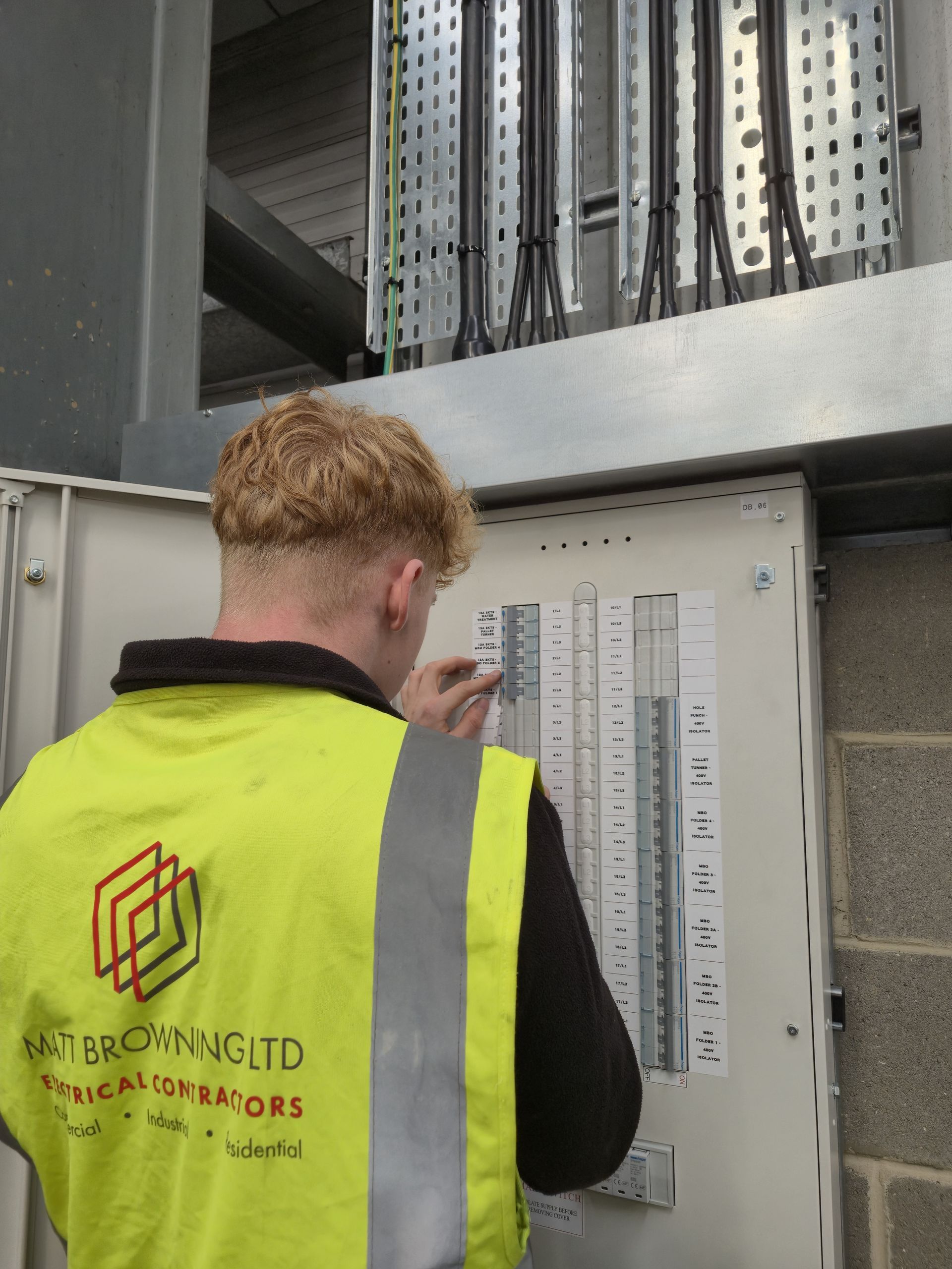 An electrical apprentice from Matt Browning Electrical is fitting circuit labelling and BS7671 circuit schedules to a 3-phase distribution board at a commercial industrial building as part of a fit-out project in Berkshire. The picture shows neatly glanded SWA cables, galvanised steel cable tray and cable trunking containment above the Hager 125A TPN distribution board.