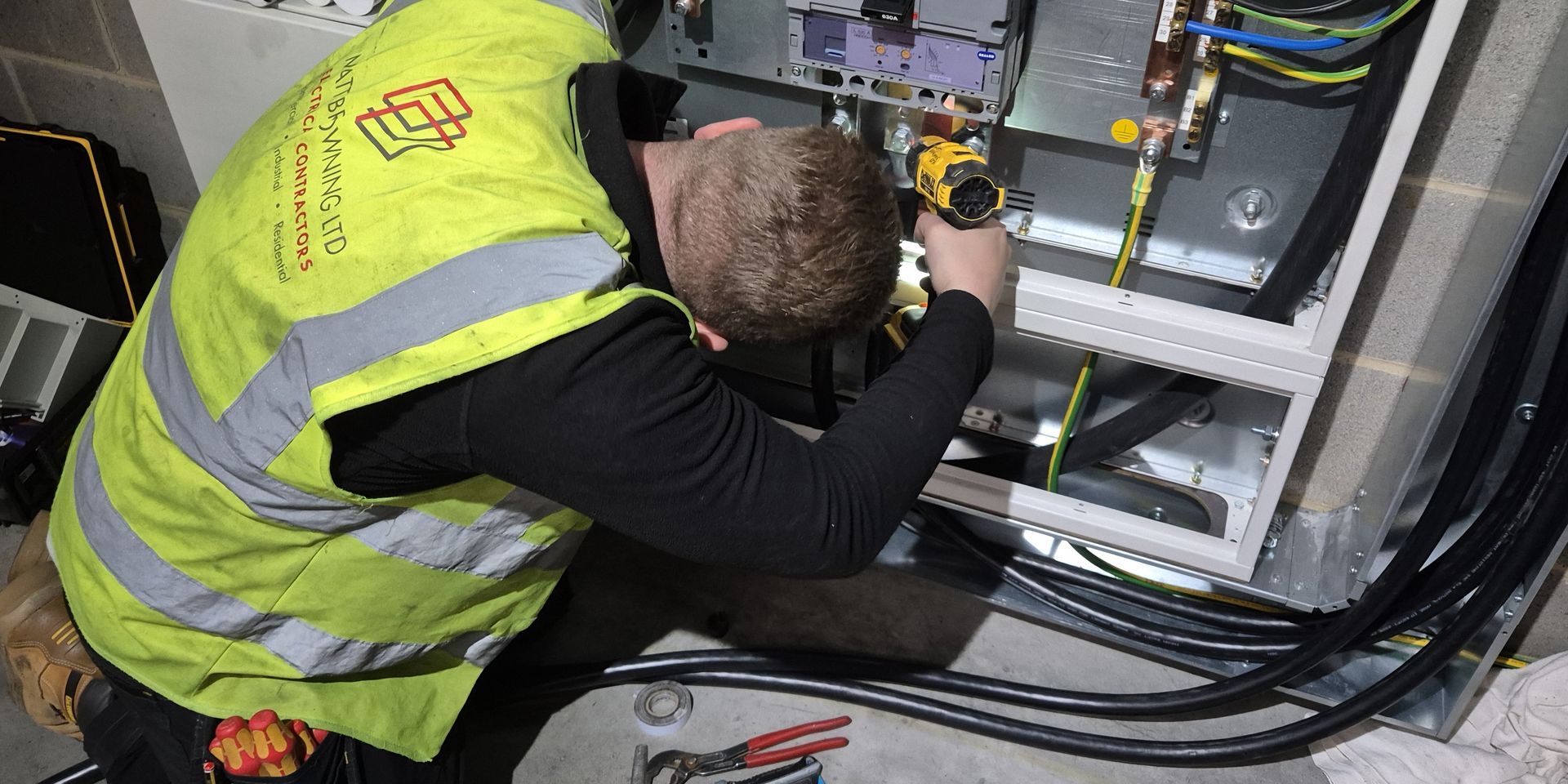An approved industrial electrician is installing new meter tails in galvanised steel trunking to feed a 630A 3-phase Hager panel board as part of a supply upgrade and commercial fit-out project near Slough, Reading, and Bracknell. The electrician is wearing PPE and a Matt Browning Electrical Contractors branded Hi-vis vest, while fitting heat shrink to crimped tri-rated cable connections.