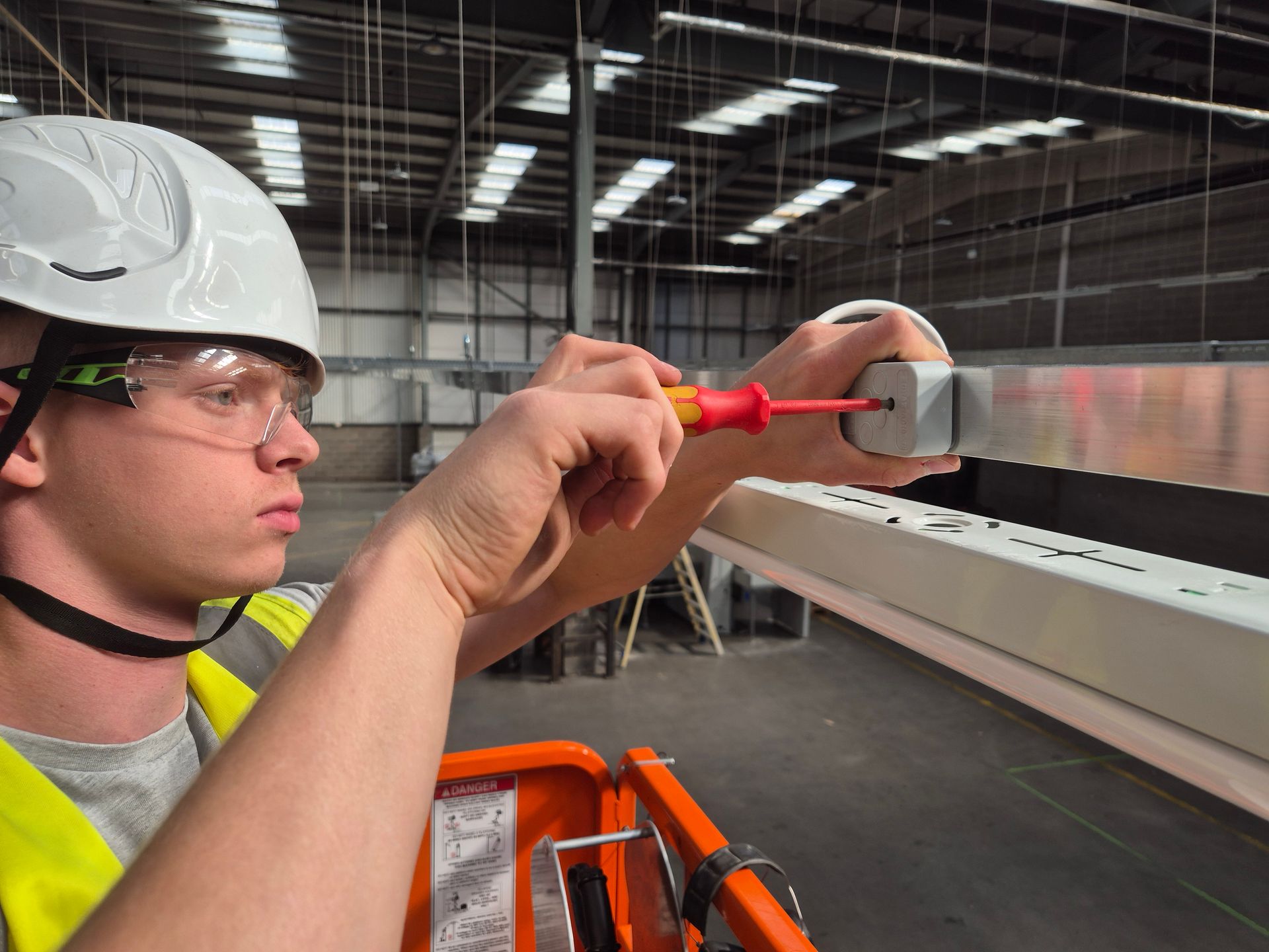An apprentice electrician fitting LED lighting battens on busbar trunking in a commercial industrial warehouse and factory area as part of an electrical fit out project by Matt Browning Electrical Contractors based near Bracknell, Reading, and Slough. The electrician is wearing PPE and NICEIC approved contractor logo on his Hi-vis vest.