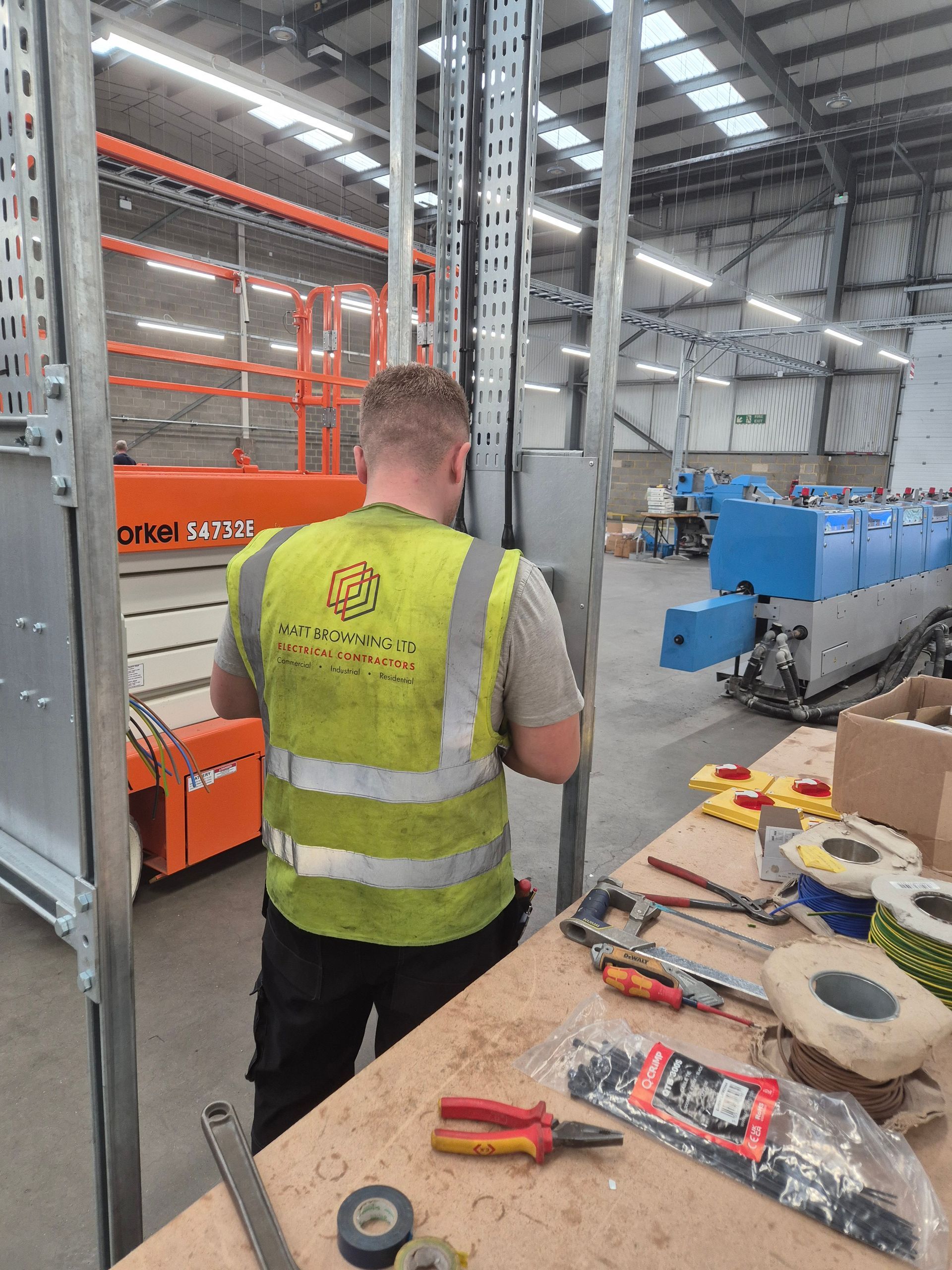 An electrician connecting and terminating cables into industrial sockets and isolators mounted on a unistrut channel frame and cable tray assembly in a factory fit out of a commercial building near Maidenhead and Reading. The electrician is wearing a branded Hi-vis vest as a member of the team from Matt Browning Electrical Contractors. The sockets and isolators will supply 3-phase power to machines and small power. All designed, installed, and electrically tested by the NICEIC approved electrician.