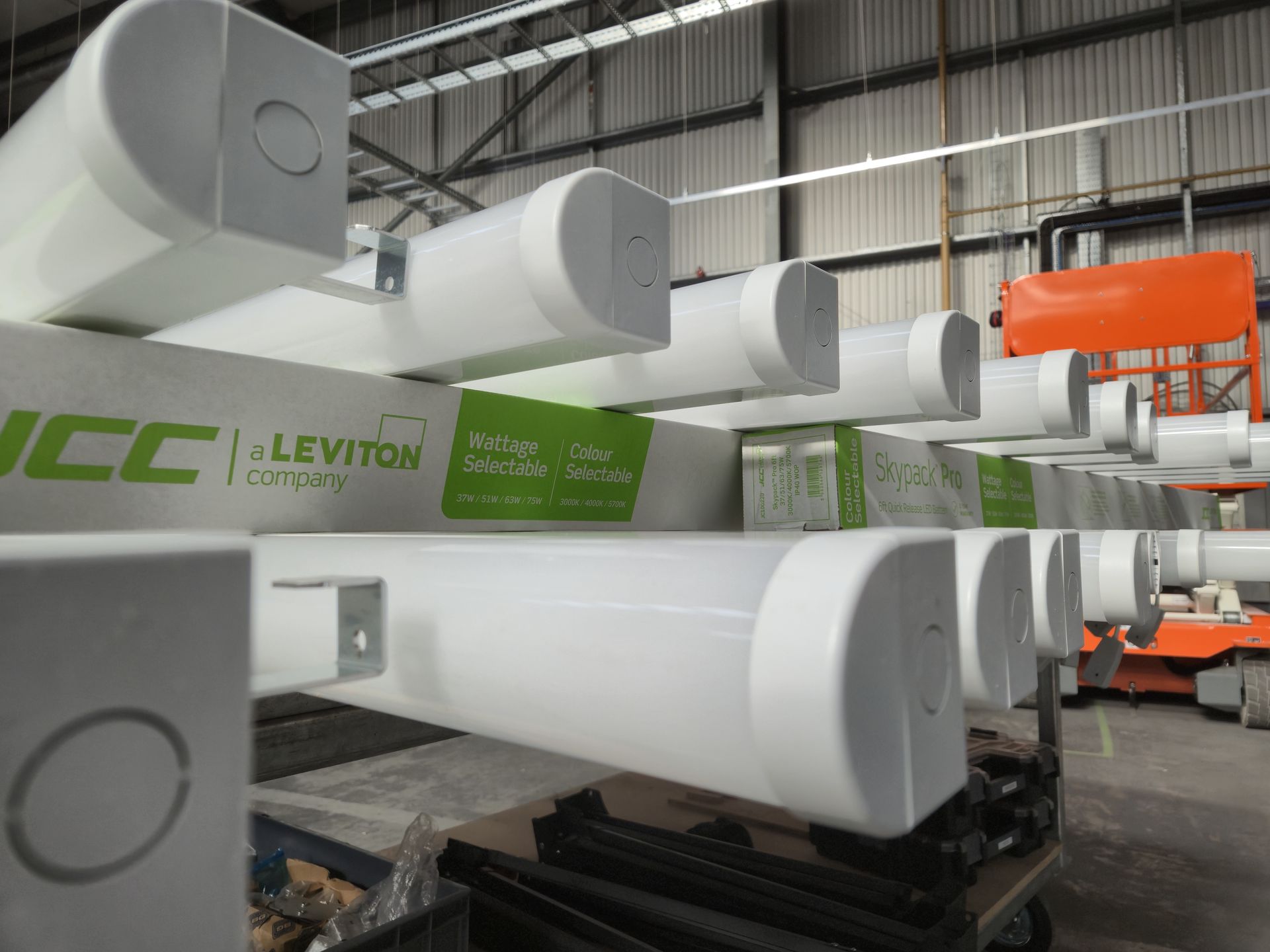 A workbench loaded with JCC Skypack LED Batten lights being prepared and installed inside a commercial building near Reading, with a scissor lift in the background to help the electricians from Matt Browning Electrical work safely at height. Some of the lighting is fitted with emergency lighting packs to ensure safe evacuation in the event of power failure in the factory.