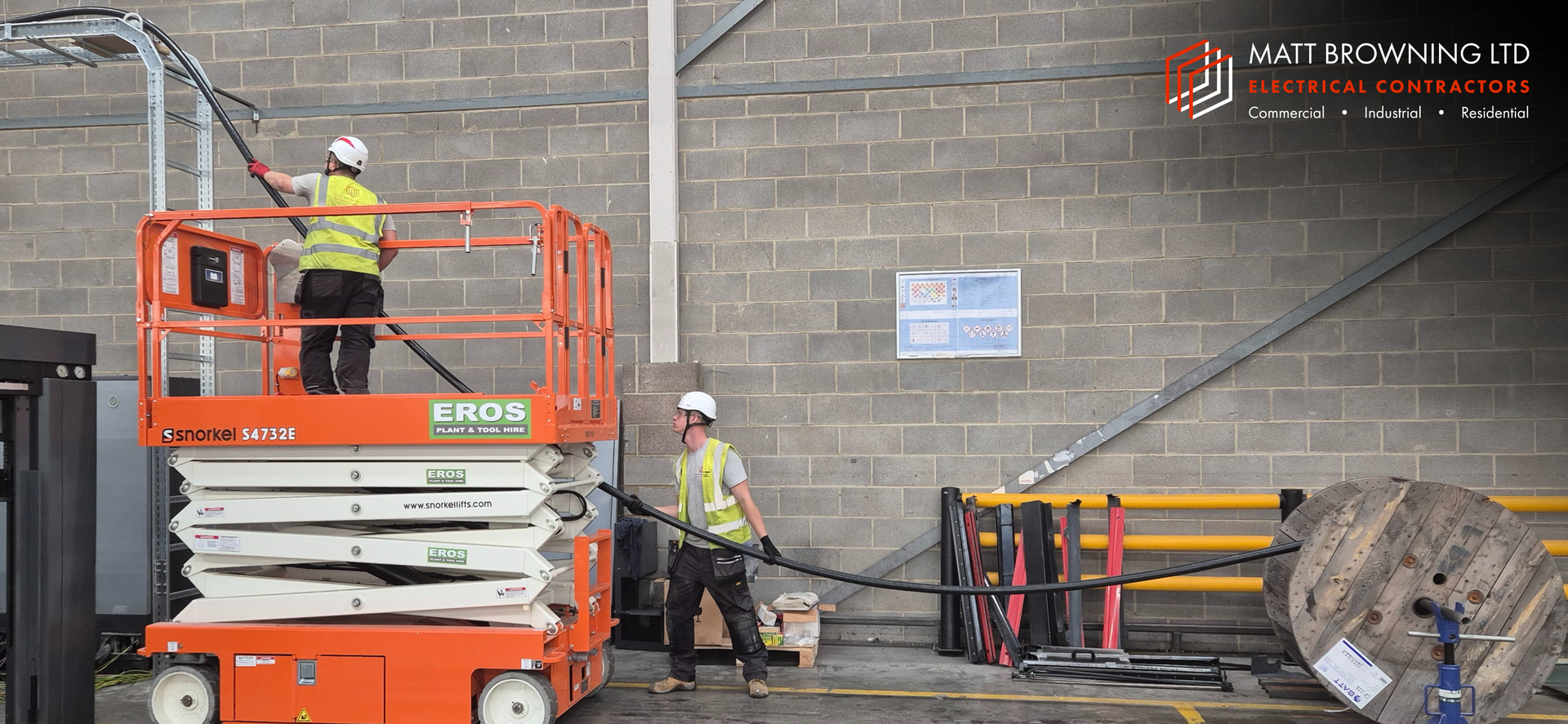 Two electricians from Matt Browning Electrical wearing Hi-vis and PPE are installing industrial cabling using a scissor lift on hire from Eros Tool & Plant Hire in a commercial industrial building near Slough Berkshire. They are pulling SWA cable onto a cable ladder system to feed a 200A 3-phase supply for an industrial machine. This work was designed and quoted as part of a fixed package for a business relocation and factory fitout by the friendly NICEIC Approved Contractor team at Matt Browning Electrical in Wokingham.